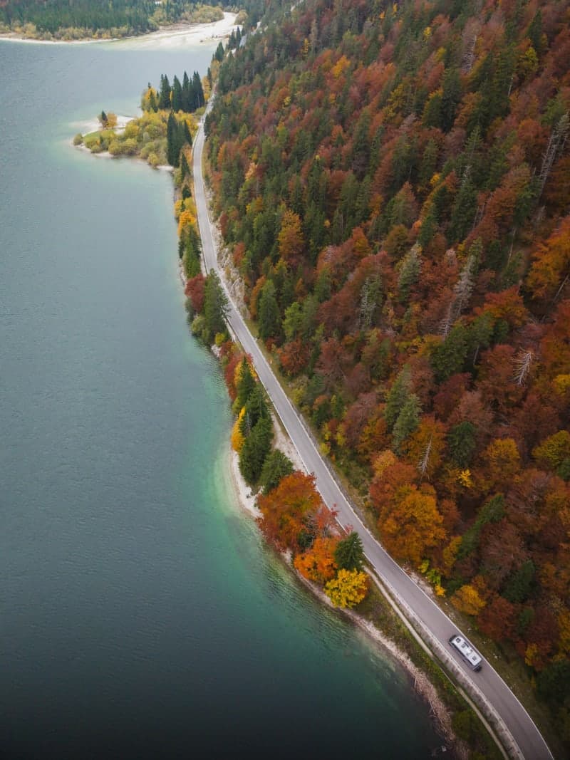 Aerial view of peak autumn maple forest with a winding road and a lake in golden October light