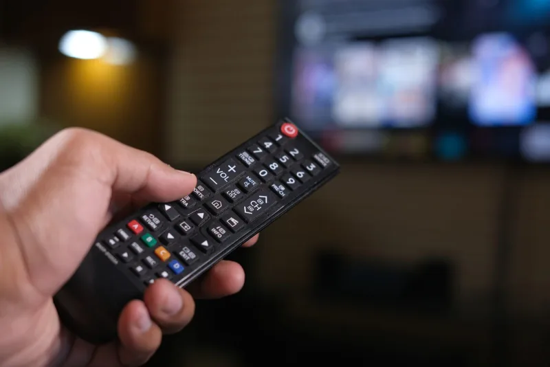 A hand holding a TV remote control with a blurred television screen behind it